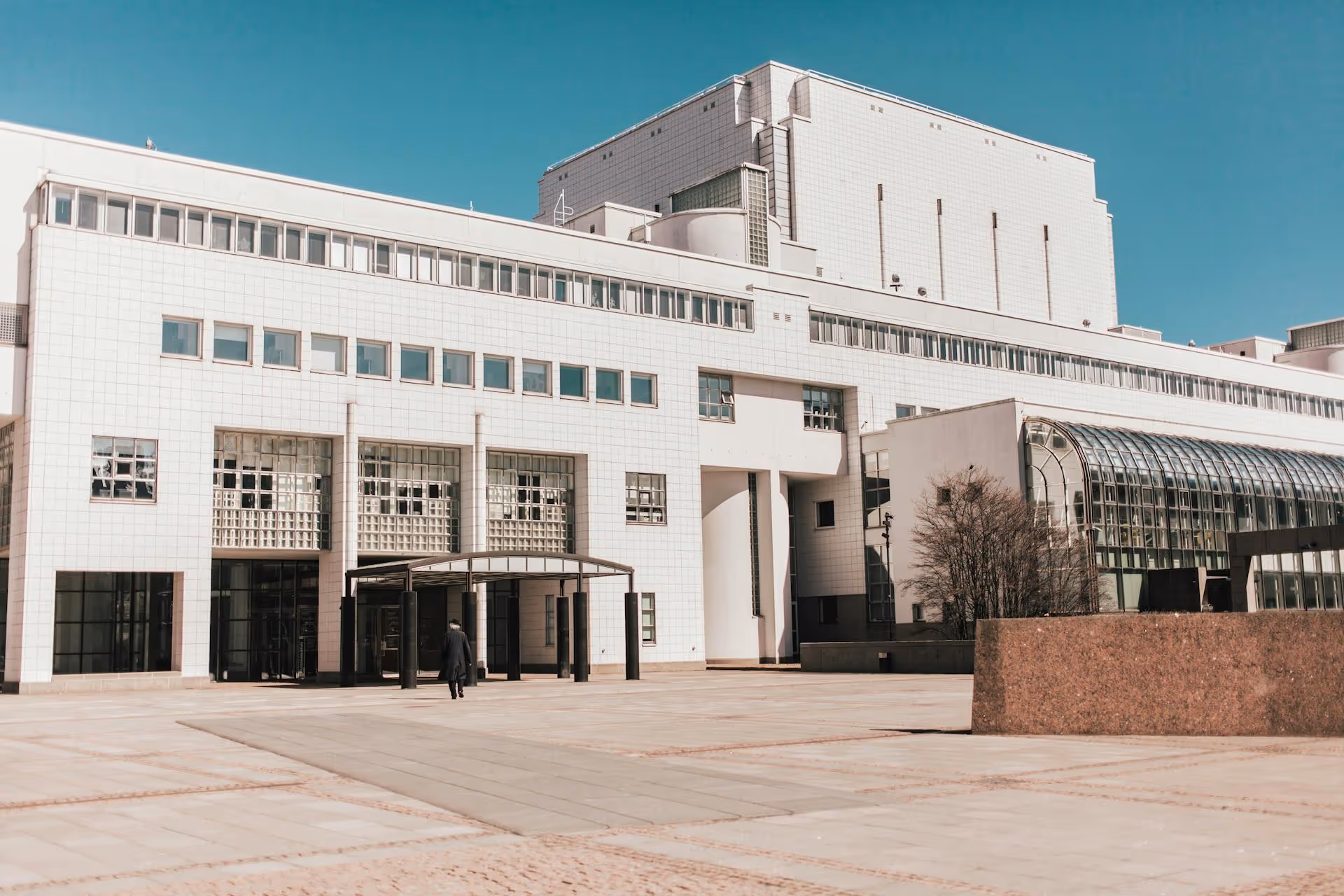 Large modern white building with rows of windows and a glass-covered entrance under a clear blue sky.
